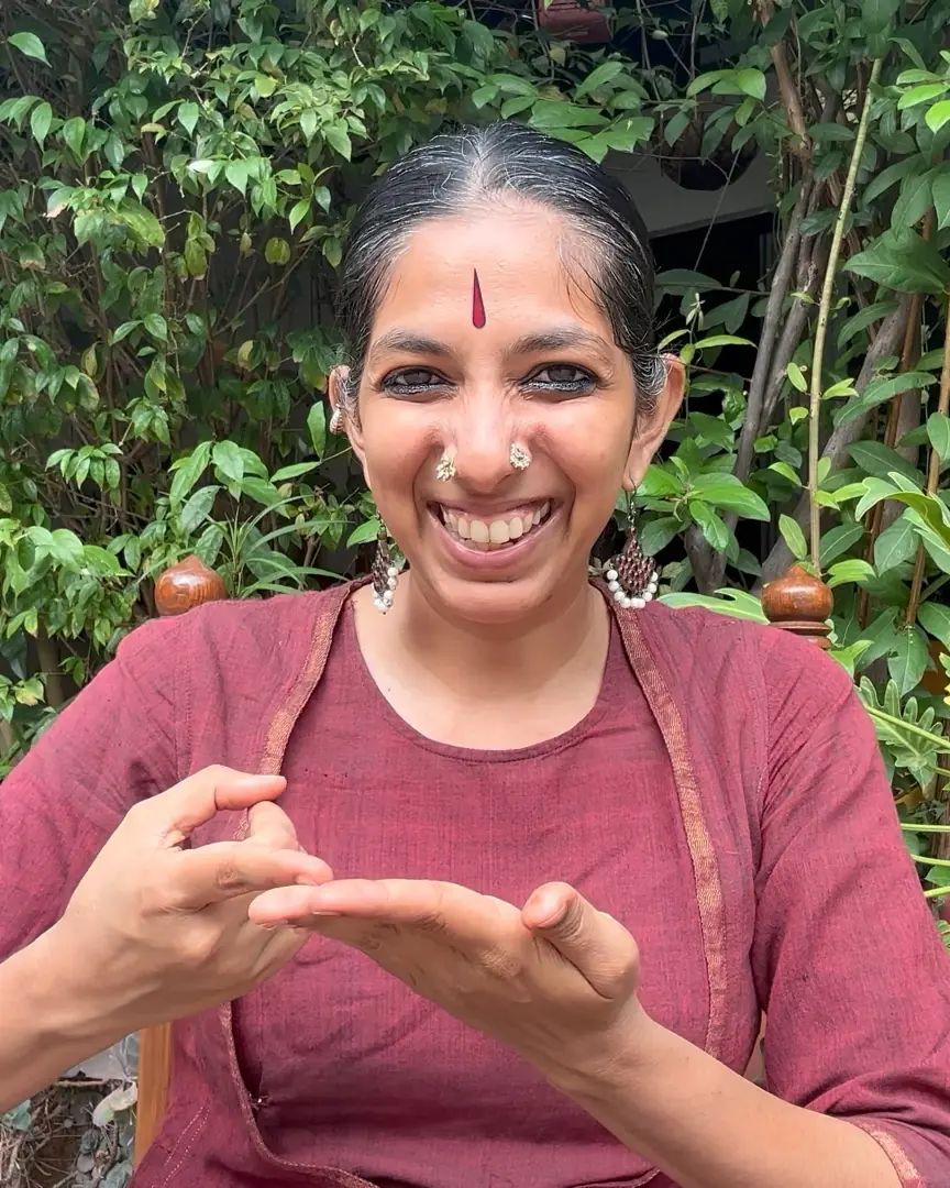 Priya in a maroon kurta, laughing, seated among garden plants with her hands in a mudra.