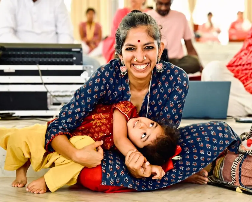 Priya on a hall floor, grinning, with her daughter Devi laid across her lap