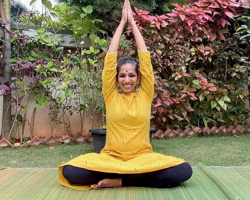 Priya on a yoga mat in a garden, arms raised with palms joined, smiling